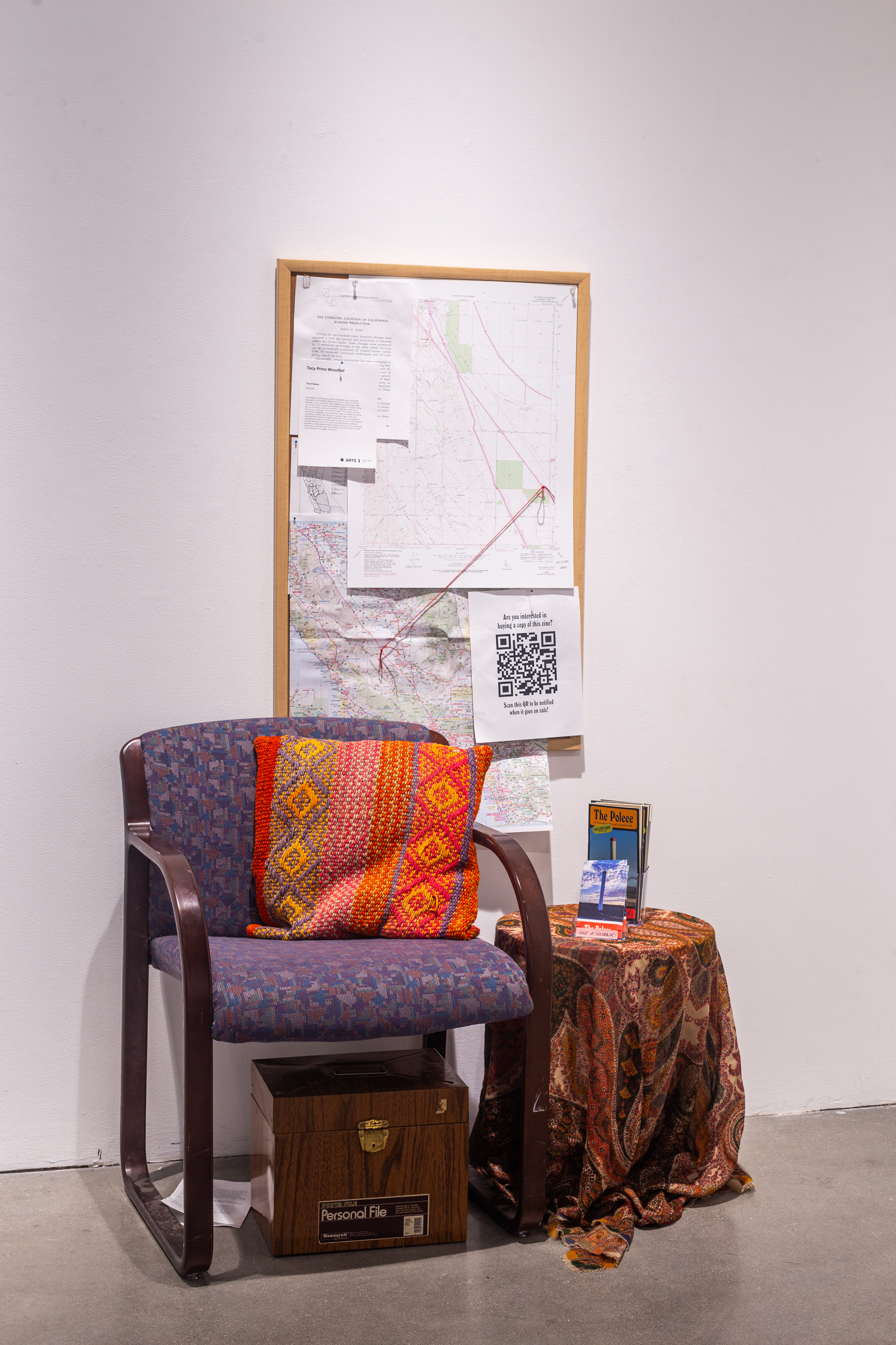 A chair sits in the center of the frame, with a pillow on it. On the right of the chair is a small circular coffee table with brochures. In the back is a white wall with a corkboard with maps and diagrams.